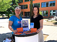 Zwei Frauen stehen an einem Messestand und halten Postkarten mit der Aufschrift "Praktikumswoche" in der Hand.